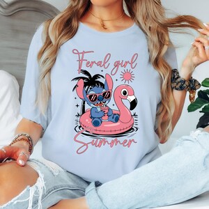 Pu&ograve; includere: T-shirt azzurra con un disegno di Stitch di Lilo & Stitch con occhiali da sole su un galleggiante a forma di fenicottero rosa. Il testo dice "Feral girl Summer" in rosa. Un design estivo e divertente.