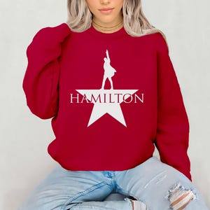Op de afbeelding: Rood sweatshirt met een wit silhouet van een persoon die op een witte ster staat met het woord "HAMILTON" eronder gedrukt.