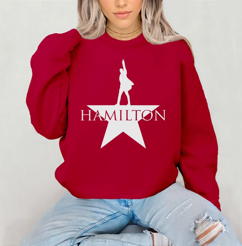 Op de afbeelding: Rood sweatshirt met een wit silhouet van een persoon die op een witte ster staat met het woord "HAMILTON" eronder gedrukt.