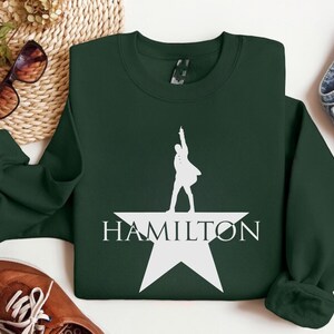 Op de afbeelding: Een donkergroen sweatshirt met een wit silhouet van een persoon die op een witte ster staat met het woord "HAMILTON" eronder.