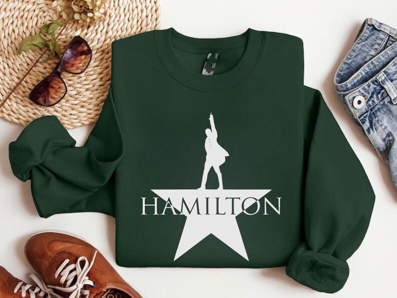 Op de afbeelding: Een donkergroen sweatshirt met een wit silhouet van een persoon die op een witte ster staat met het woord "HAMILTON" eronder.