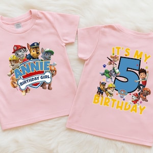 May include: Two light pink short-sleeved t-shirts featuring Paw Patrol characters. The left shirt displays the text "ANNIE BIRTHDAY GIRL" in a blue and red shield. The right shirt features the text "IT'S MY 5 BIRTHDAY" with a large blue number 5.