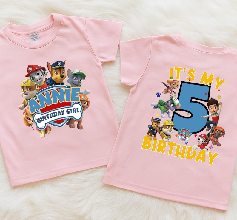 May include: Two light pink short-sleeved t-shirts featuring Paw Patrol characters. The left shirt displays the text "ANNIE BIRTHDAY GIRL" in a blue and red shield. The right shirt features the text "IT'S MY 5 BIRTHDAY" with a large blue number 5.