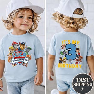 May include: A light blue t-shirt featuring Paw Patrol characters. The front displays the text 'LUCA BIRTHDAY BOY' with a shield graphic. The back shows a large blue number '3' with the text 'IT'S MY BIRTHDAY' and additional character graphics.