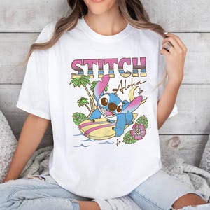 Puede incluir: Camiseta blanca con un gr&aacute;fico colorido del personaje Stitch surfeando en una tabla de surf. La palabra "STITCH" est&aacute; impresa encima del personaje, con "Aloha" escrito debajo. El dise&ntilde;o tambi&eacute;n incluye palmeras y flores.