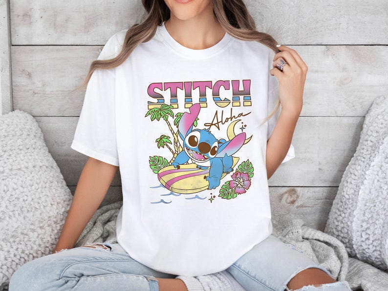 Puede incluir: Camiseta blanca con un gr&aacute;fico colorido del personaje Stitch surfeando en una tabla de surf. La palabra "STITCH" est&aacute; impresa encima del personaje, con "Aloha" escrito debajo. El dise&ntilde;o tambi&eacute;n incluye palmeras y flores.