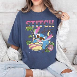 Puede incluir: Una camiseta azul marino con la palabra "STITCH" en una fuente retro colorida. El gr&aacute;fico incluye un personaje de dibujos animados en una tabla de surf, palmeras, una luna creciente y la palabra "Aloha".