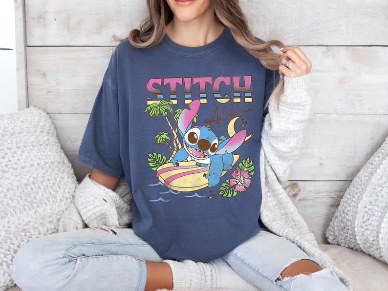 Puede incluir: Una camiseta azul marino con la palabra "STITCH" en una fuente retro colorida. El gr&aacute;fico incluye un personaje de dibujos animados en una tabla de surf, palmeras, una luna creciente y la palabra "Aloha".