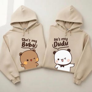 May include: Two beige hoodies with matching designs. One says "She's my Bubu" with a brown bear illustration. The other says "He's my Dudu" with a white panda illustration. Both hoodies have drawstrings and a hood.