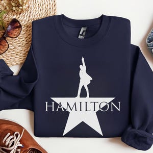 Op de afbeelding: Een donkerblauw sweatshirt met een wit silhouet van een persoon die op een witte ster staat met het woord "HAMILTON" eronder.