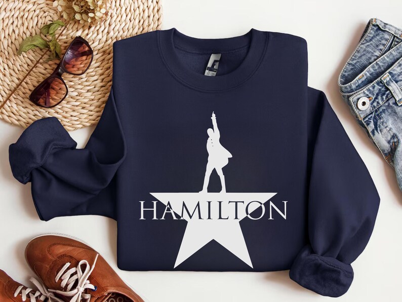 Op de afbeelding: Een donkerblauw sweatshirt met een wit silhouet van een persoon die op een witte ster staat met het woord "HAMILTON" eronder.