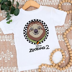 May include: White short-sleeved t-shirt featuring a cartoon sloth character from Disney's Zootopia 2. The sloth is inside a black and white checkered circle. The text "Zootopia 2" is printed below the sloth image in black.