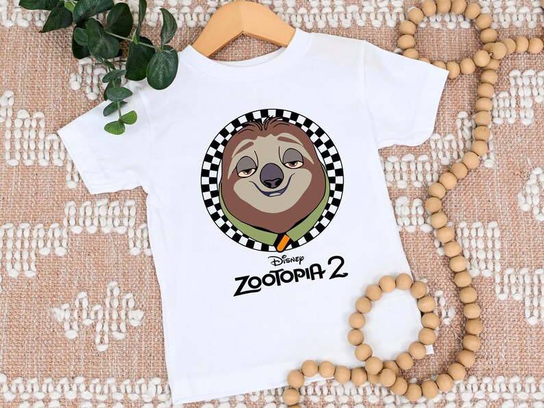 May include: White short-sleeved t-shirt featuring a cartoon sloth character from Disney's Zootopia 2. The sloth is inside a black and white checkered circle. The text "Zootopia 2" is printed below the sloth image in black.