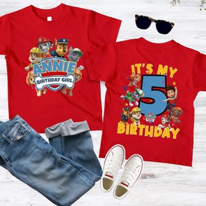 May include: Two red t-shirts featuring Paw Patrol characters. One shirt displays the text "ANNIE BIRTHDAY GIRL" with a shield graphic. The other shirt features a large blue number "5" with the text "IT'S MY BIRTHDAY" and various characters.