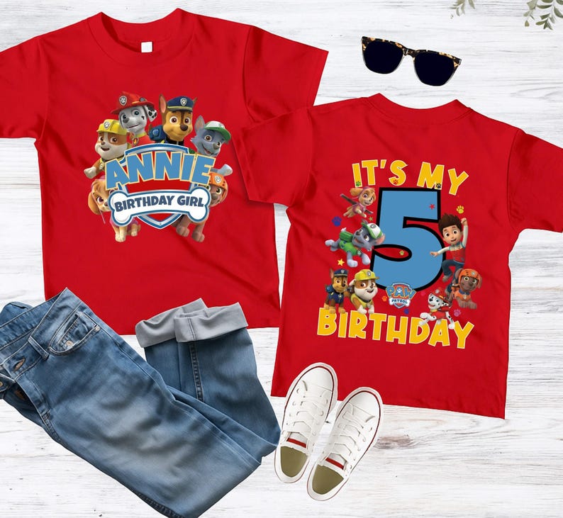 May include: Two red t-shirts featuring Paw Patrol characters. One shirt displays the text "ANNIE BIRTHDAY GIRL" with a shield graphic. The other shirt features a large blue number "5" with the text "IT'S MY BIRTHDAY" and various characters.