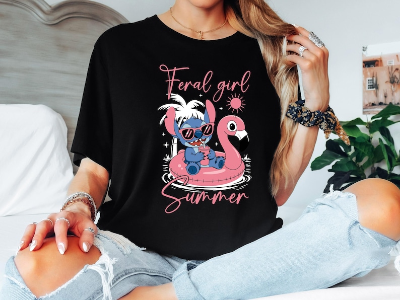 Pu&ograve; includere: T-shirt nera con un personaggio dei cartoni animati con occhiali da sole, che galleggia su un galleggiante a forma di fenicottero rosa. Il testo "Feral girl Summer" &egrave; scritto in rosa. Il design include un sole, palme e stelle.