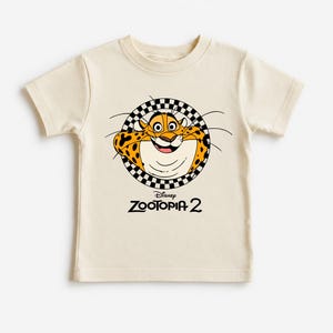 May include: Cream-colored t-shirt featuring a cartoon cheetah character from Disney's Zootopia 2. The cheetah is inside a black and white checkered circle. The text "Zootopia 2" is below the cheetah.