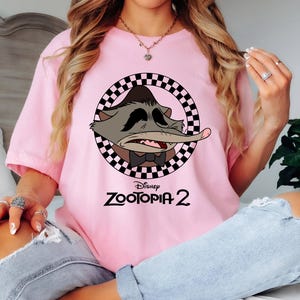 May include: A light pink t-shirt featuring a cartoon character from Disney's Zootopia 2. The character is inside a black and white checkered circle. The text "Disney Zootopia 2" is printed below the character in black.