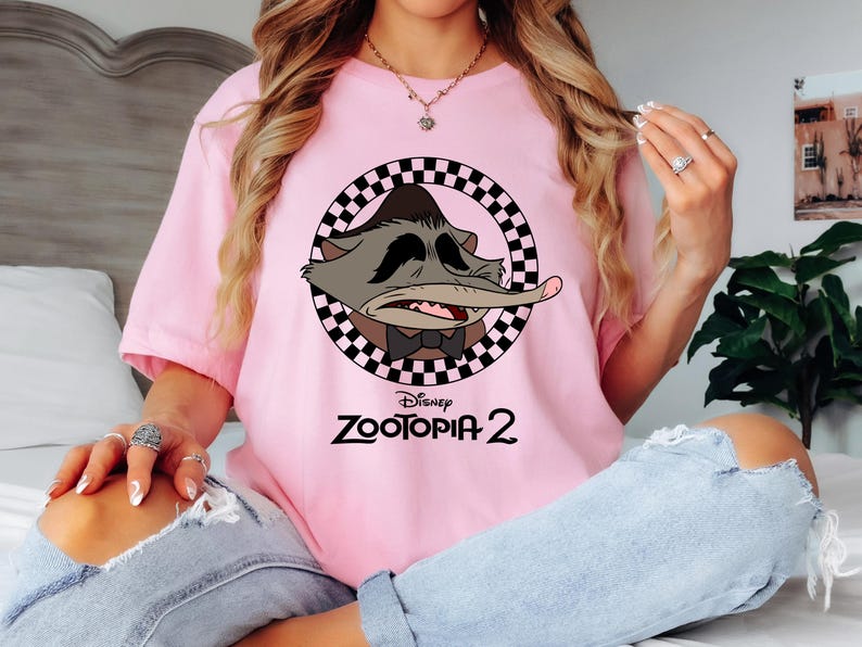 May include: A light pink t-shirt featuring a cartoon character from Disney's Zootopia 2. The character is inside a black and white checkered circle. The text "Disney Zootopia 2" is printed below the character in black.