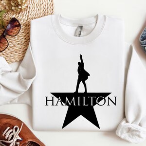 Op de afbeelding: Wit sweatshirt met een zwart silhouet van een persoon die op een ster staat met het woord "HAMILTON" eronder.