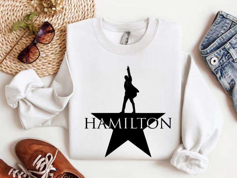 Op de afbeelding: Wit sweatshirt met een zwart silhouet van een persoon die op een ster staat met het woord "HAMILTON" eronder.