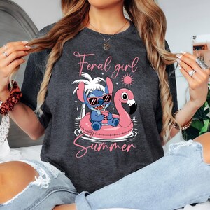 Pu&ograve; includere: T-shirt grigio scuro con un personaggio dei cartoni animati con occhiali da sole su un galleggiante a forma di fenicottero rosa. Il testo "Feral girl Summer" &egrave; scritto in rosa sopra e sotto la grafica. Un design estivo casual.