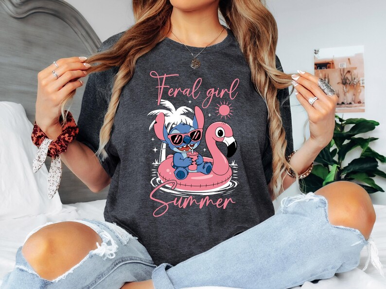 Pu&ograve; includere: T-shirt grigio scuro con un personaggio dei cartoni animati con occhiali da sole su un galleggiante a forma di fenicottero rosa. Il testo "Feral girl Summer" &egrave; scritto in rosa sopra e sotto la grafica. Un design estivo casual.