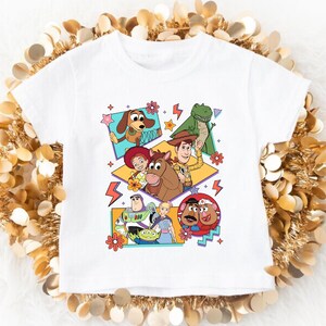 May include: White t-shirt featuring a colorful graphic of characters from the animated film Toy Story. The design includes Woody, Buzz Lightyear, and other characters in a retro style. The shirt is made of a soft material.