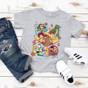 May include: A gray t-shirt featuring a colorful graphic of characters from the animated film Toy Story. The shirt is paired with blue denim jeans and white sneakers with black stripes. A small potted plant is also visible.