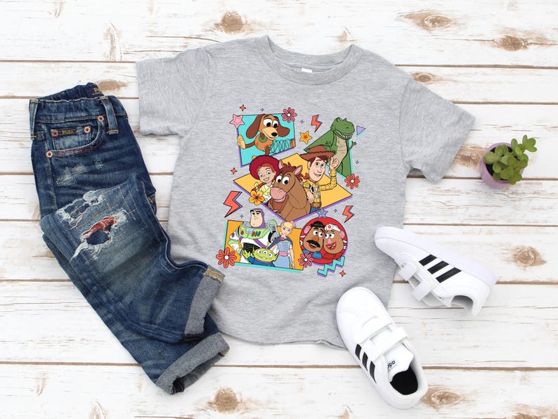 May include: A gray t-shirt featuring a colorful graphic of characters from the animated film Toy Story. The shirt is paired with blue denim jeans and white sneakers with black stripes. A small potted plant is also visible.