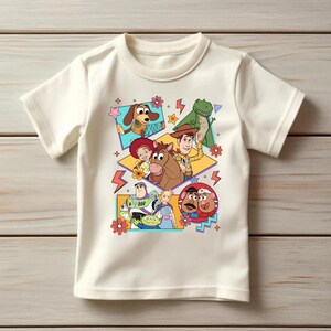 May include: Off-white short-sleeved t-shirt featuring a colorful graphic of characters from the animated film Toy Story. The design includes images of Woody, Buzz Lightyear, and other characters in a playful arrangement.