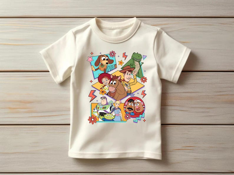 May include: Off-white short-sleeved t-shirt featuring a colorful graphic of characters from the animated film Toy Story. The design includes images of Woody, Buzz Lightyear, and other characters in a playful arrangement.
