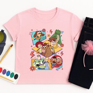 May include: A light pink t-shirt featuring a colorful graphic of characters from the Toy Story movie. The image is surrounded by a pair of dark blue jeans, a pink headband, and a pair of navy blue shoes. A watercolor paint set and brush are also included.