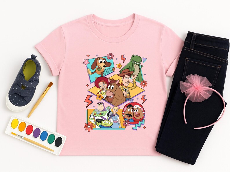 May include: A light pink t-shirt featuring a colorful graphic of characters from the Toy Story movie. The image is surrounded by a pair of dark blue jeans, a pink headband, and a pair of navy blue shoes. A watercolor paint set and brush are also included.
