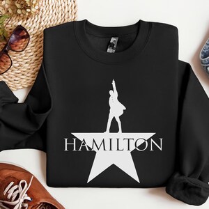 Op de afbeelding: Zwart sweatshirt met een wit silhouet van een persoon die op een witte ster staat met het woord "HAMILTON" eronder.