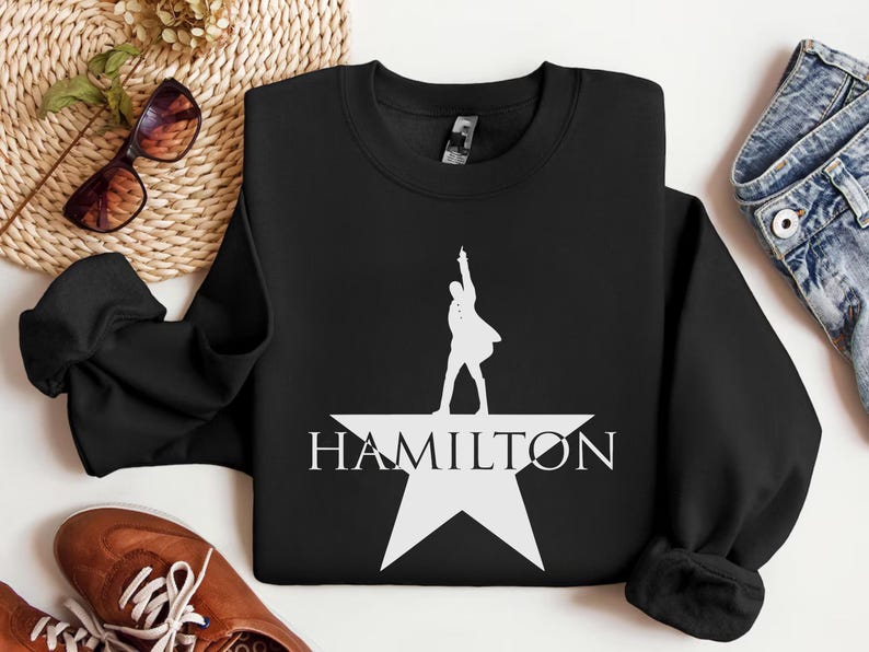 Op de afbeelding: Zwart sweatshirt met een wit silhouet van een persoon die op een witte ster staat met het woord "HAMILTON" eronder.