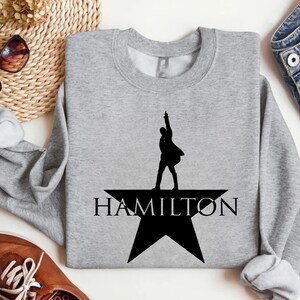Op de afbeelding: Een grijs sweatshirt met een zwart silhouet van een persoon die op een ster staat met het woord "HAMILTON" eronder.