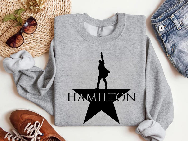 Op de afbeelding: Een grijs sweatshirt met een zwart silhouet van een persoon die op een ster staat met het woord "HAMILTON" eronder.