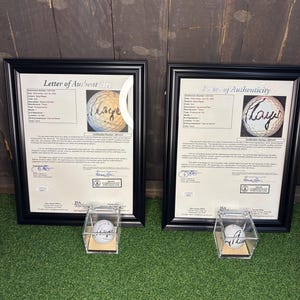 May include: Two framed "Letter of Authenticity" documents with black frames, each featuring a golf ball with a signature. Below each frame is a clear display case holding a golf ball. The background is green turf and a dark wood wall.