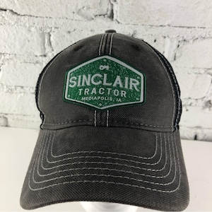 May include: A gray baseball cap with a mesh back and a curved brim. The front features a green and white patch with the text "SINCLAIR TRACTOR MEDIAPOLIS, IA". White stitching accents the cap's seams.
