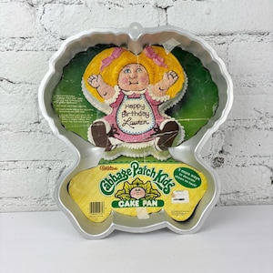 Puede incluir: Molde para pastel Cabbage Patch Kids de color plateado. El molde tiene la forma de una muñeca Cabbage Patch Kids con cabello amarillo y un vestido rosa. Las palabras "Happy Birthday Lauren" están escritas en el vestido. El molde también tiene las palabras "Cabbage Patch Kids Cake Pan".