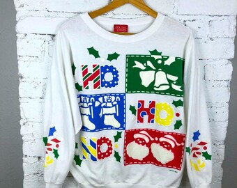 May include: White long-sleeved sweatshirt with a colorful holiday design. The design includes the words "HO HO HO" in various colors and festive images like bells, stockings, and Santa hats. The sweatshirt has a crew neck and is hanging on a wooden hanger.