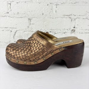 Op de afbeelding: Bronzen klompen met een geweven leren bovenwerk en een donkerbruine houten plateauzool. De klompen hebben een ronde neus en bronzen studs rond de zool. De hakhoogte is ongeveer 7.6 cm. Gouden binnenzool.