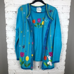 May include: A turquoise blue cardigan sweater set with a matching top. The cardigan has a floral design with colorful flowers, butterflies, and stars. The top has a floral design with colorful flowers. The set is made of a soft, comfortable material.