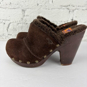 Op de afbeelding: Bruine suède klompen met een imitatiebontvoering en houten hakken. De klompen hebben een ronde neus, messingkleurige studs en een logo aan de zijkant. De hakken zijn ongeveer 10 cm hoog.