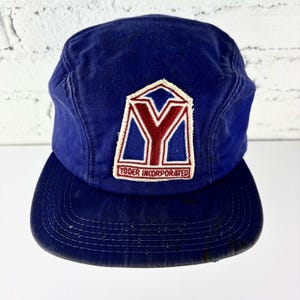 Può includere: Un cappellino da baseball blu con visiera piatta. Il cappellino presenta un logo bianco, rosso e blu con la scritta "Yoder Incorporated". Il cappellino è realizzato in un tessuto resistente e ha uno stile classico e vintage.