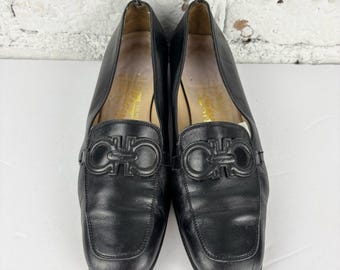 May include: Pair of black leather loafers with a square toe and a decorative buckle. The shoes have a low heel and a classic design. The interior of the shoe has gold lettering.