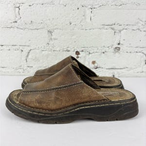 May include: A pair of brown leather slide sandals with a distressed appearance. The sandals have a thick, black sole and visible white stitching. The open-toe design and slip-on style suggest casual footwear.