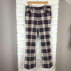 Puede incluir: Un pantalón a cuadros de estilo vintage. El pantalón presenta una base color crema con un patrón de cuadros negro, rojo y azul. El pantalón está colgado de una percha de madera contra una pared de ladrillos blancos.