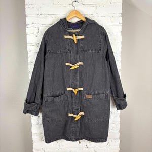 May include: A dark gray denim hooded coat with four large front pockets and wooden toggle closures. The coat is hanging on a wooden hanger against a white brick wall.